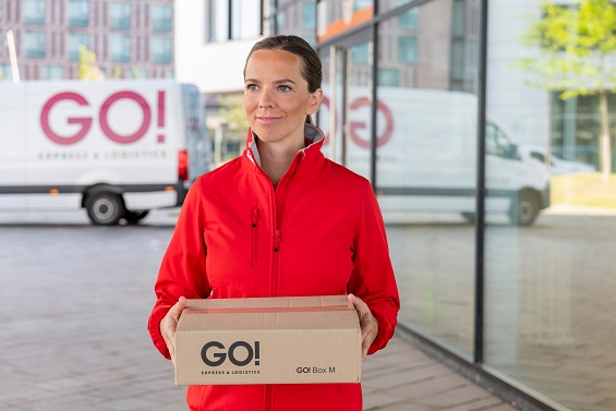 Lächelnde GO! Kurierin mit kleinem GO! Paket in der Hand vor einem Bürogebäude